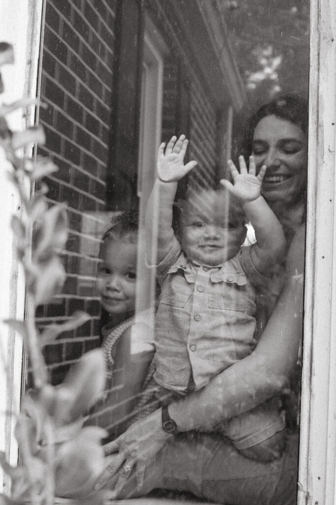 Black and white photo of mom with toddler and baby looking through window during in-home family session in Atlanta, GA